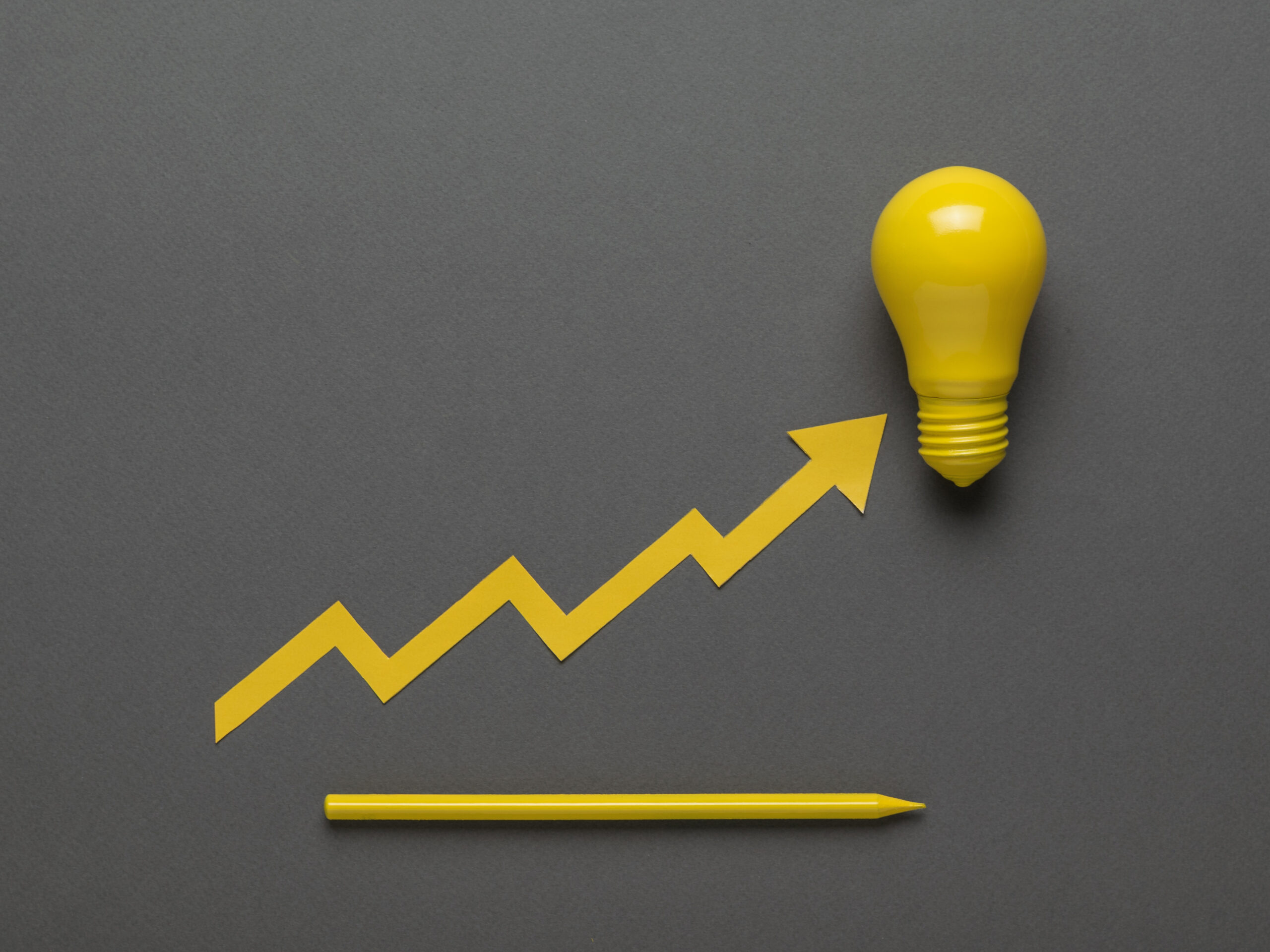 A yellow ascending curve resting on a yellow light bulb and a yellow pencil on a dark gray background. The concept of business and ideas.